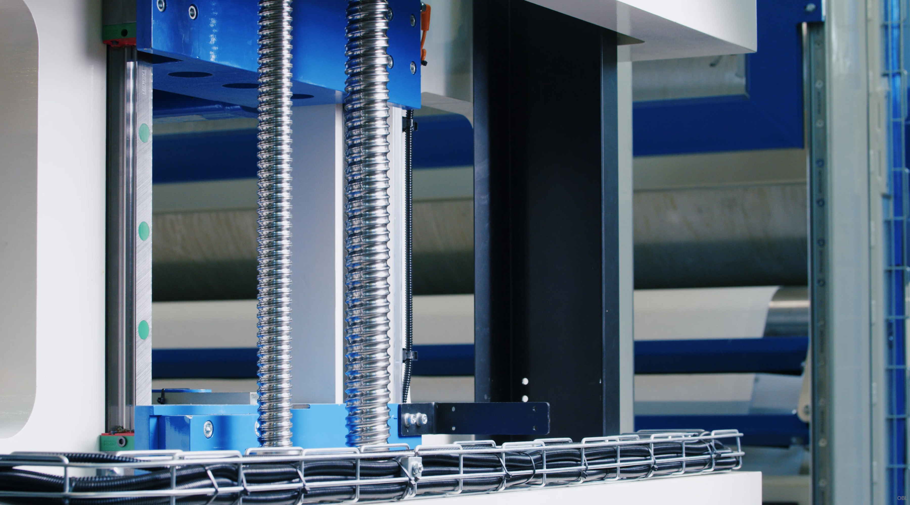 The subject of the photo is an industrial mechanism known as an 'eshuttle' machine which appears to be composed primarily of metallic components with a blue hue, suggesting it might include materials like steel or aluminum for its construction. The image captures various parts such as metal rods and supports in shades ranging from silver-gray indicative of bare metal to bright blue possibly due to protective coatings or paint designed for resistance against corrosion or wear during operation within an industrial environment. This setup is part of a larger machine, potentially used in industries like manufacturing where precise positioning might be necessary, hinting at its role as a vital component involved in complex processes that require mechanical precision and robustness.